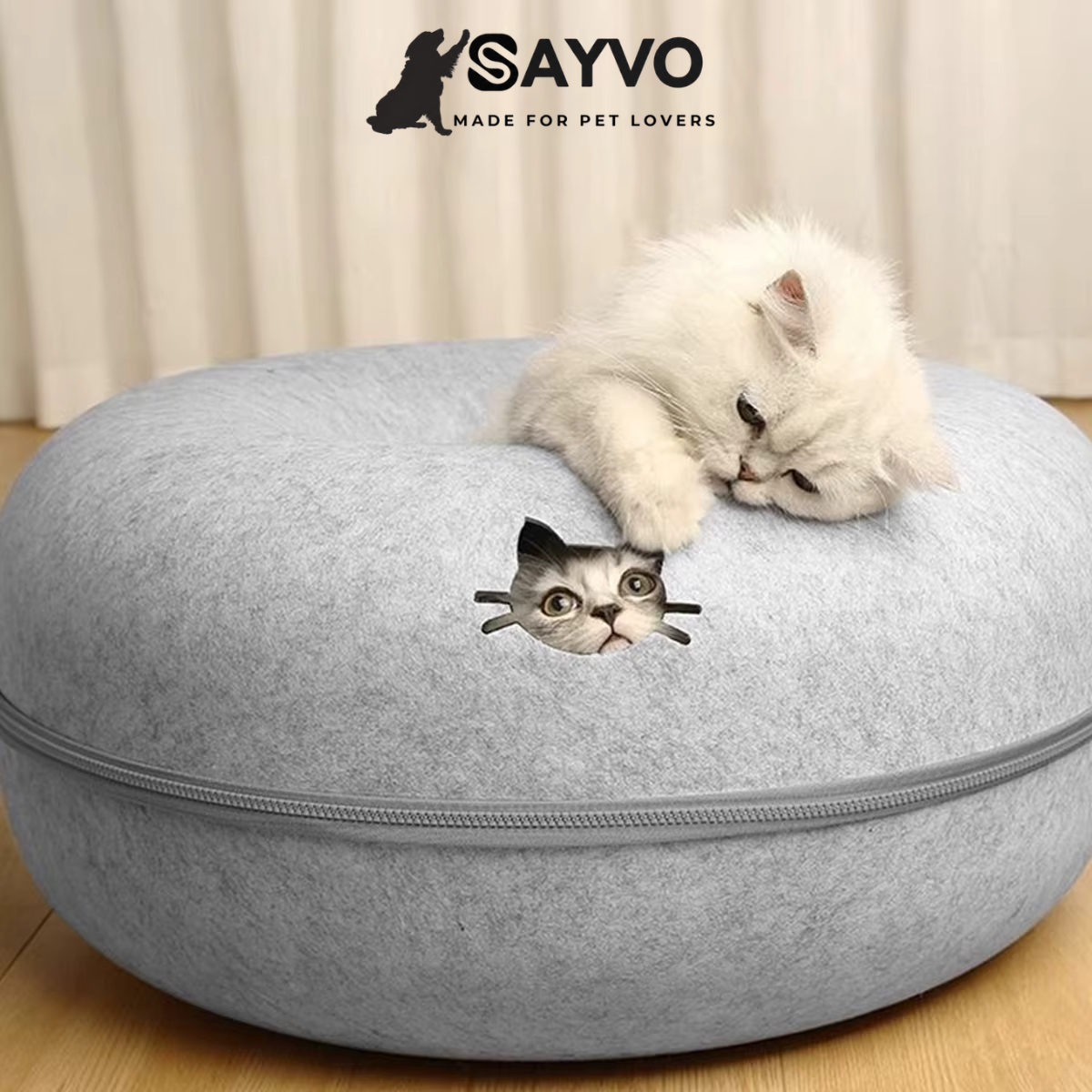 White kitten resting on a light grey round felt cat tunnel bed while another kitten peeks through the cat-shaped opening.