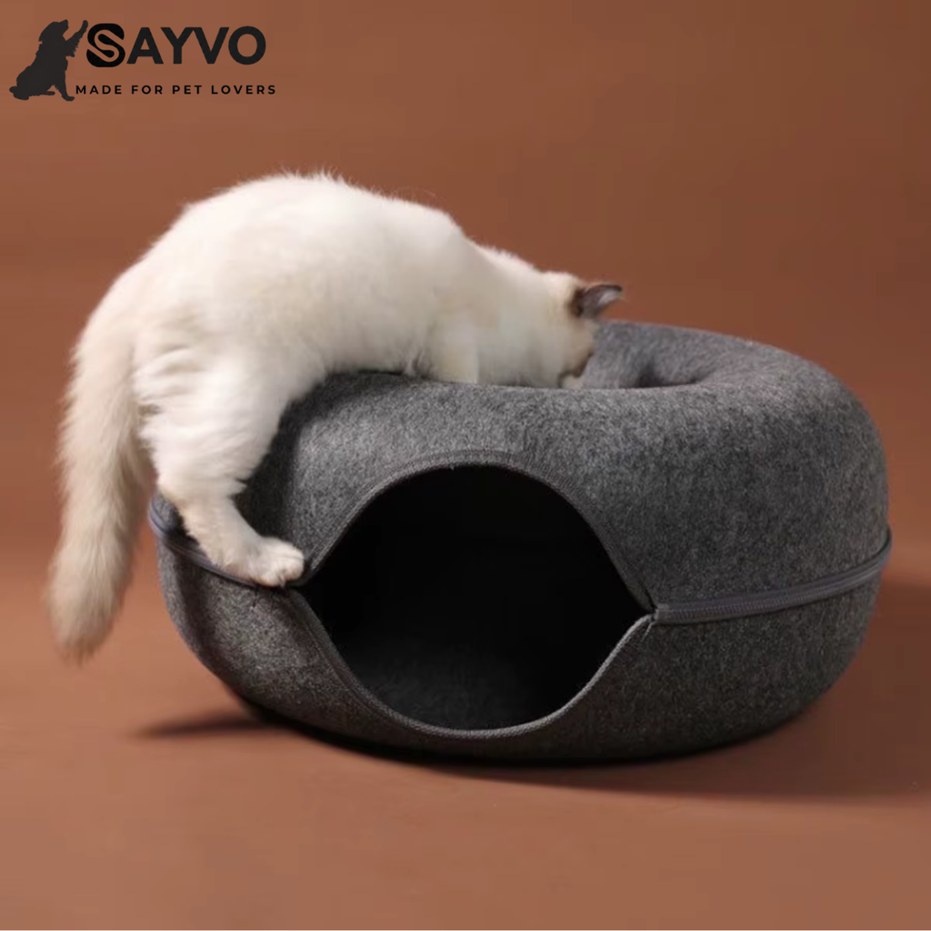 White cat exploring the central nest of a dark grey round felt cat tunnel bed with a circular opening and zippered sides.